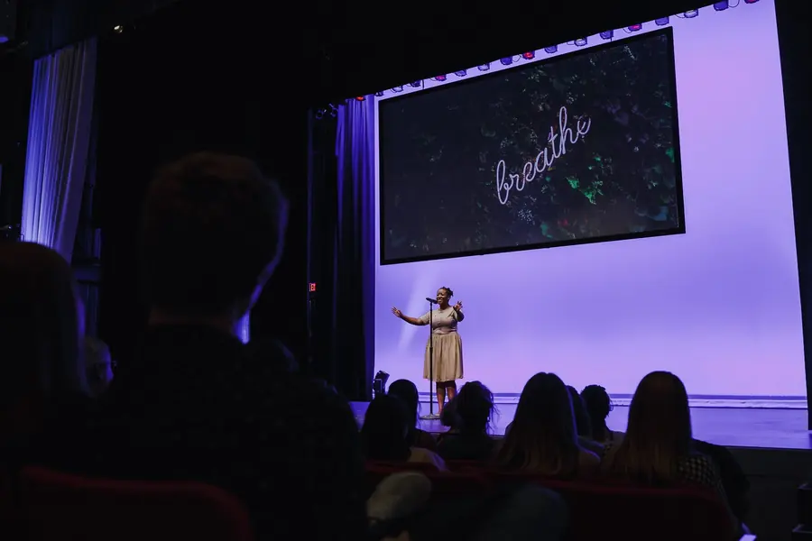 Wide view of Asha speaking on a violet-lit stage to a seated audience