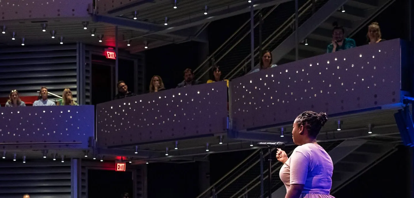 Asha Bland speaking on a theater stage to a full audience on multiple levels